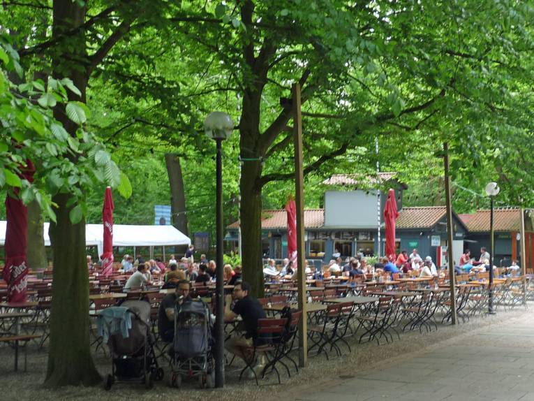 Menschen sitzen auf Bänken an Tischen unter Bäumen