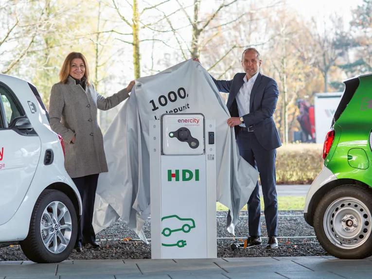 Eine Frau und ein Mann zwischen zwei Autos neben einer E-Ladesäule heben eine Abdeckung hoch
