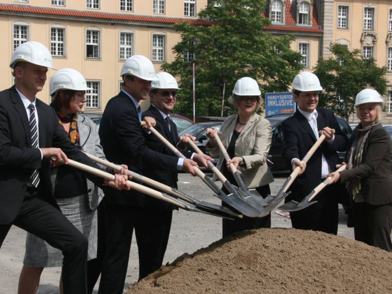 Männer und Frauen mit Helmen und Schaufeln vor einem Sandhaufen