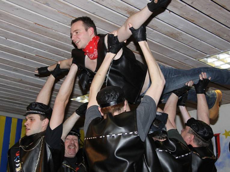 Männer mit Lederwesten und -mützen heben einen Mann in die Höhe.