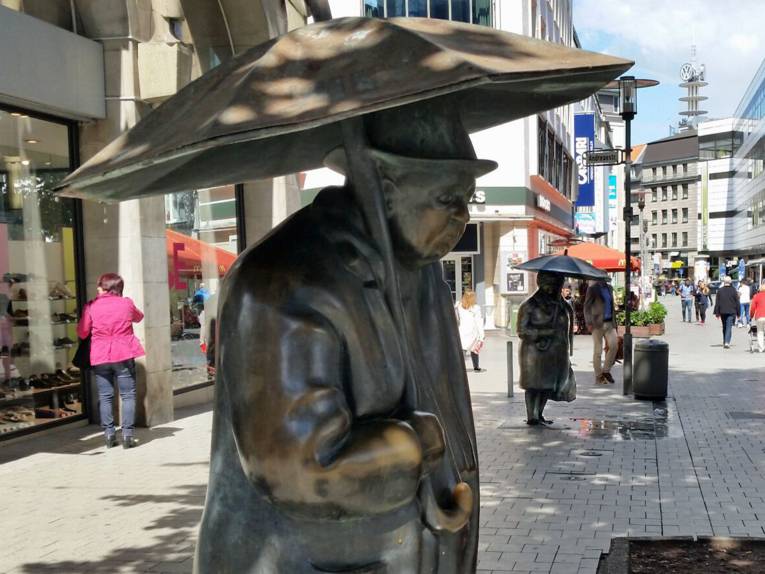 Plastiken einer Frau und eines Mannes mit Regenschirmen in einer Fußgängerzone.