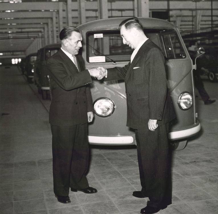 Zwei Männer schütteln sich die Hand vor einem VW Bulli. Historische Schwarz-Weiß-Aufnahme.