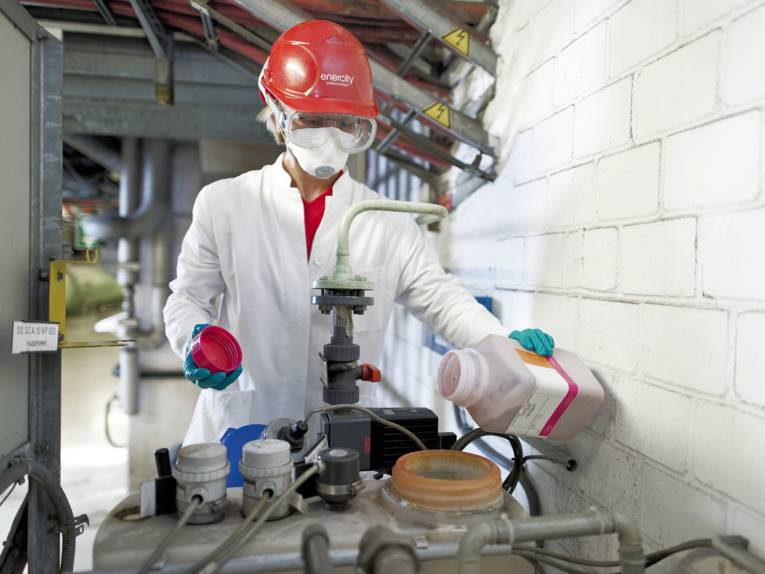 Mensch mit Schutzkleidung füllt Flüssigkeit in großen Behälter.