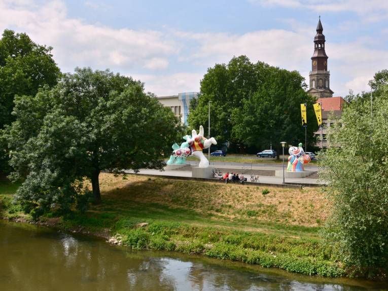 Blick über einen kleinen Fluss an dessen anderem Ufer Kunststatuen stehen.