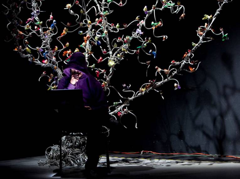 Eine Szene aus einem Stück: Ein Mann im violetten Kapuzenpulli sitzt vor einem Laptop. Hinter ihm steht ein weißer Baum voller Plastikvögel.