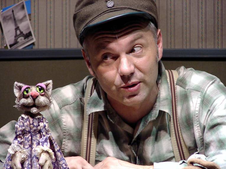 Christian Kruse spricht als Bauer verkleidet mit einer Katze im Kleid, die vor ihm auf dem Tisch sitzt.
