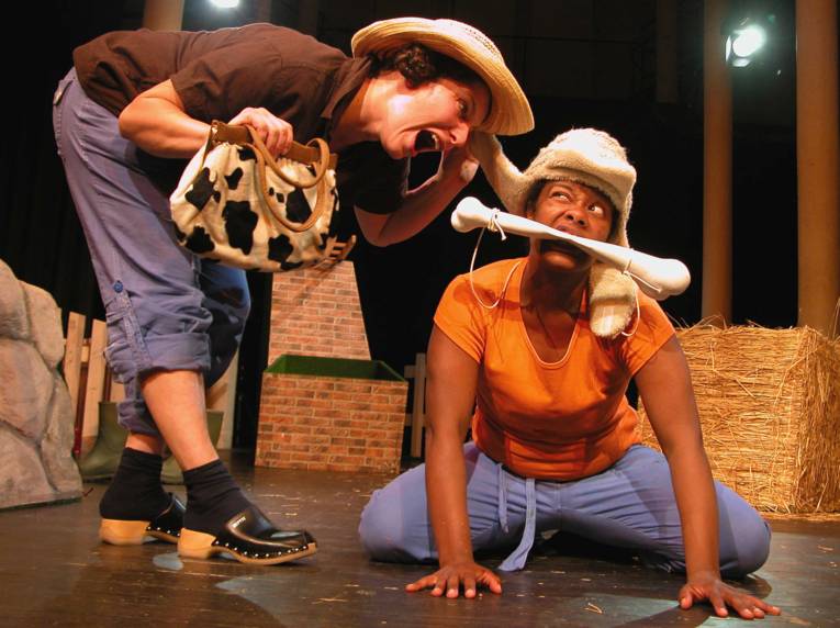 Zwei Darstellerinnen auf der Bühne, die eine hält eine gescheckte Kuhfellhandtasche in Händen, die andere hat einen großen Knochen zwischen ihren Zähnen.

