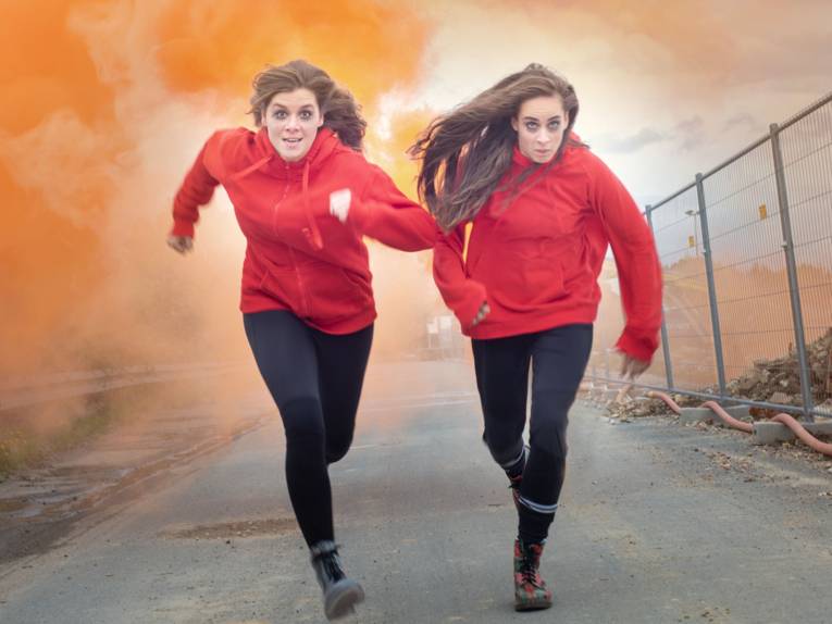 Zwei junge Frauen rennen eine Schotterstraße entlang, hinter ihnen Rauch
