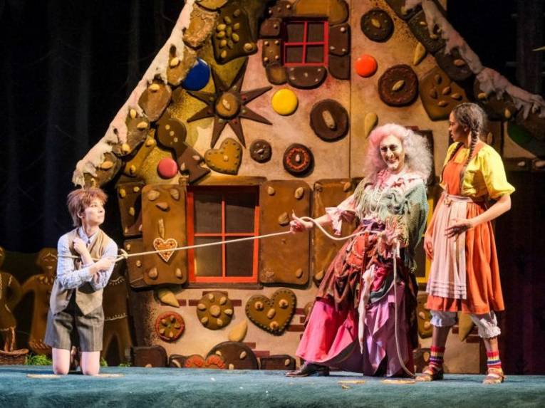 Zwei Kinder und eine Hexe vor einem großen Lebkuchenhaus.