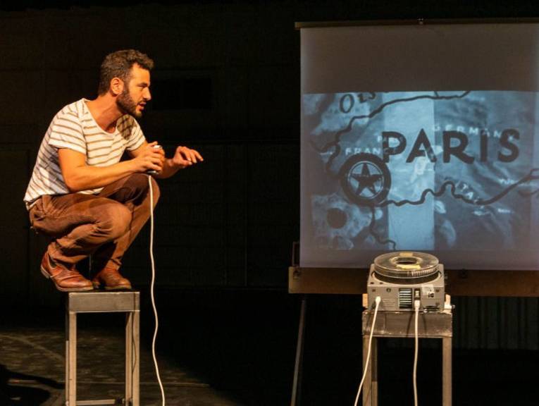 Ein Mann hockt auf einen Hocker,neben ihm ein Projektor und eine Leinwand.