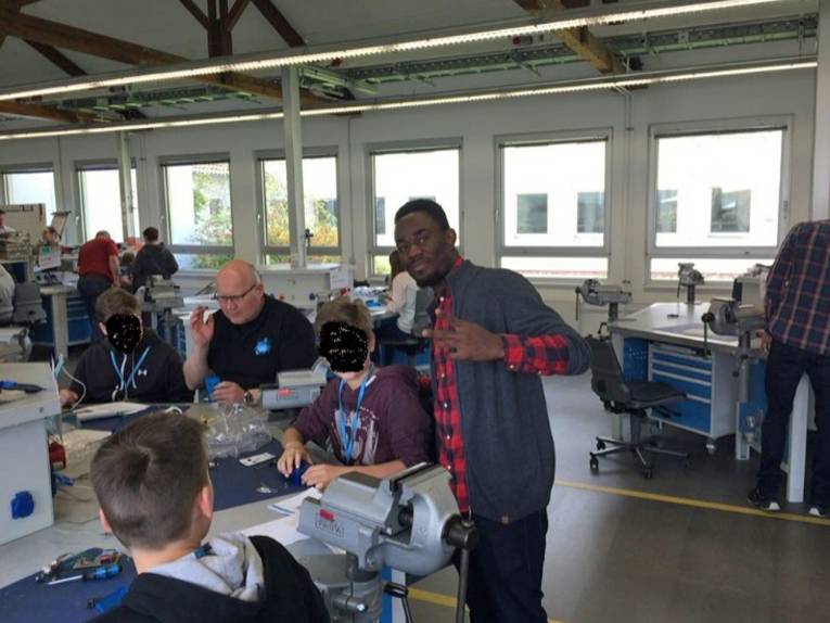 Stehender junger Mann in einer Arbeitshalle an einem Tisch, an dem drei weitere junge Männer sowie ein Ausbilder sitzen.