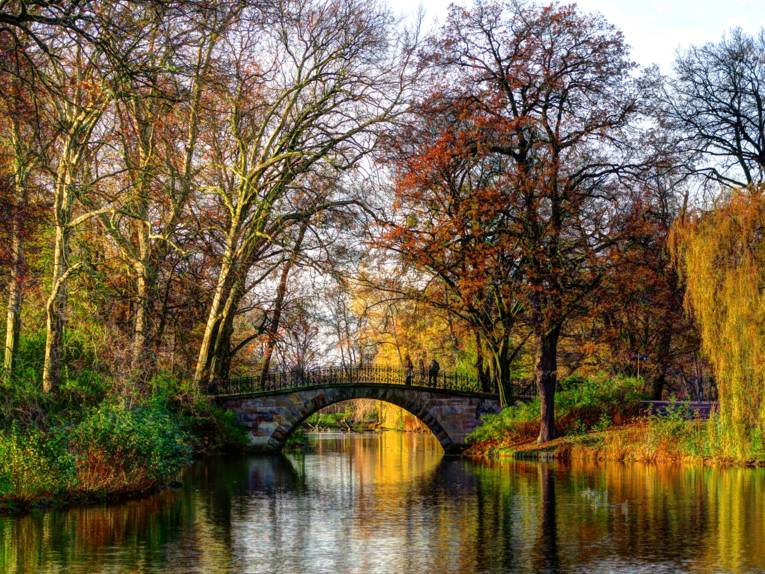 Kleine Brücke über ein Gewässer, umrahmt von Bäumen und Büschen mit verschiedenfarbenen Herbstlaub.