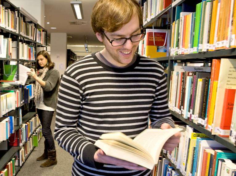 Ein junger Mann steht zwischen Regalen mit Büchern und blättert in einem Werk. Im Hintergrund ist eine junge Frau zu sehen, die ein Buch aus einem Real zieht.
