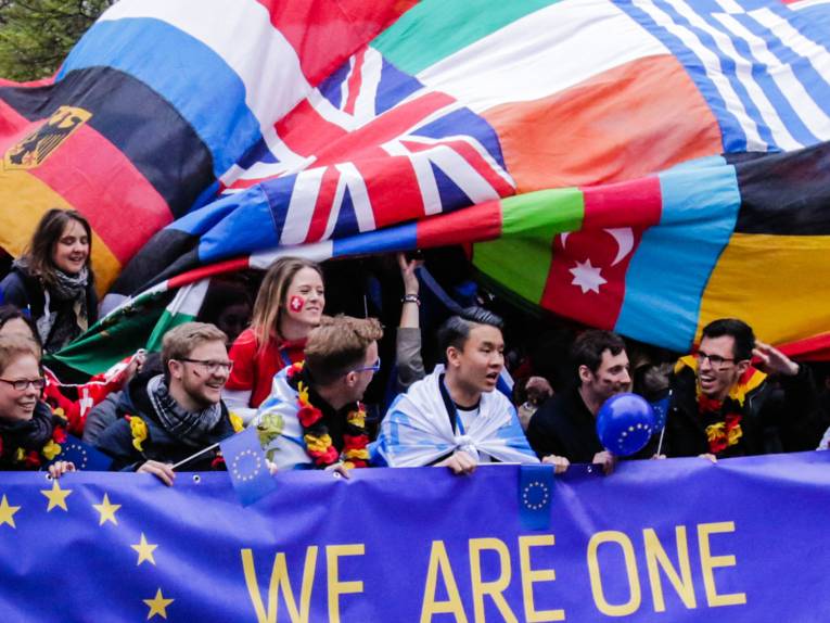 Menschen mit Flaggen im Hintergrund halten ein großes Spruchband mit Europasymbol und der Aufschrift "We are one"