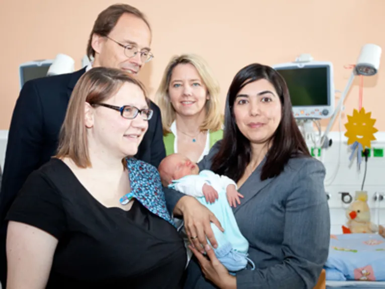 Drei Frauen und ein Mann stehen zusammen mit einem Neugeborenen in einem Krankenhauszimmer.