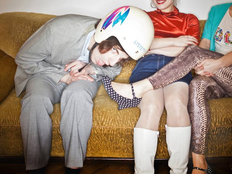 Ein junger Mann neigt sich mit seinem Kopf zum Fuß einer jungen Frau hinuter, zwischen den beiden sitzt ein weiteres Mädchen.