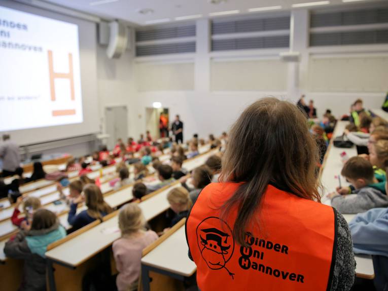 Gefüllter Hörsaal, im Vordergrund eine Frau mit einer roten Weste mit dem Aufdruck Kinder Uni Hannover