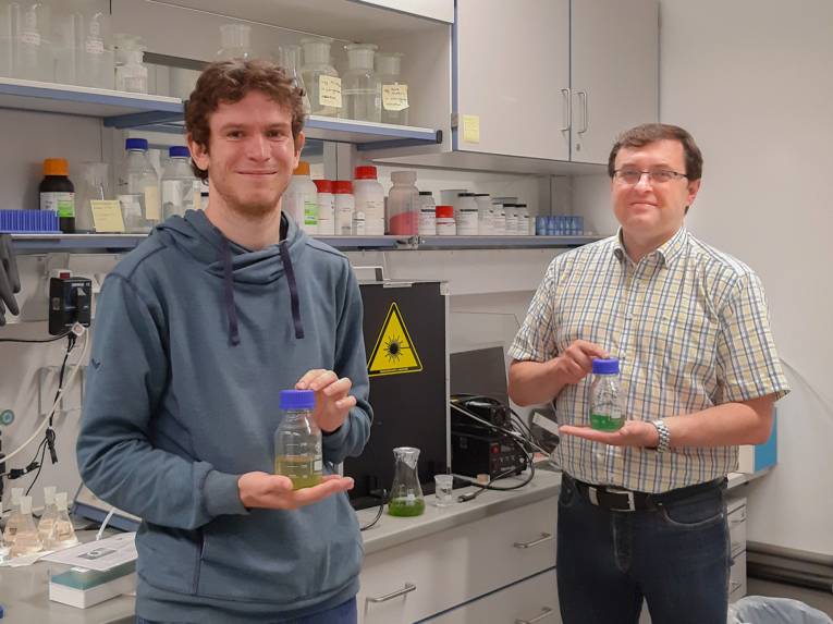 Zwei Männer in einem Labor, beide halten eine Glasflasche mit grüner Flüssigkeit in den Händen.