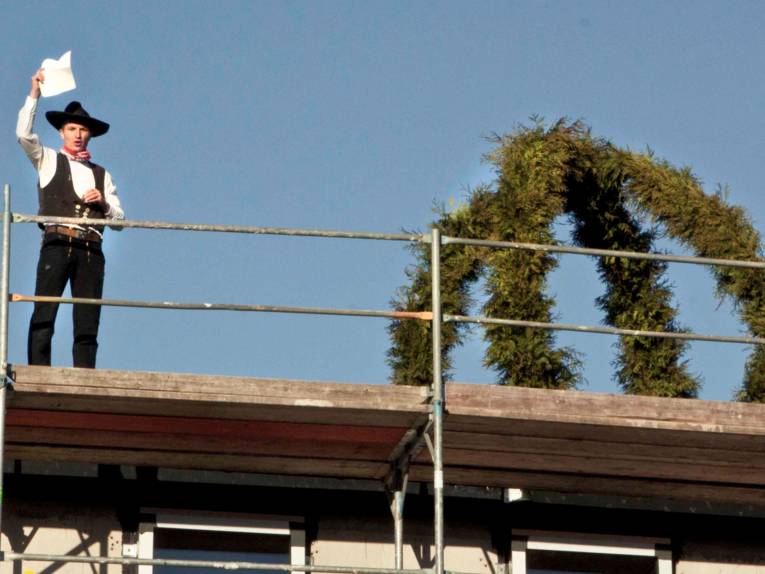 Ein Zimmermann steht neben einem Richtkranz auf einem Dach.