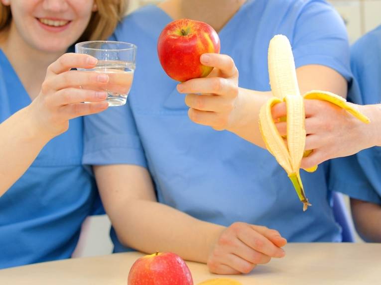 Hände halten ein Glas Wasser, einen Apfel und eine geschälte Banane.