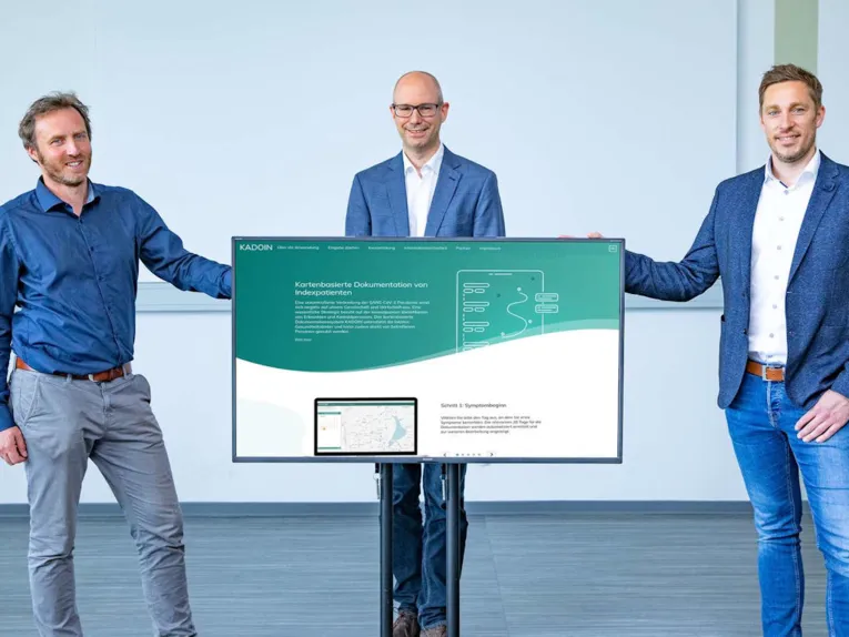 Drei Männer stehen neben und hinter einem Flachbildschirm.