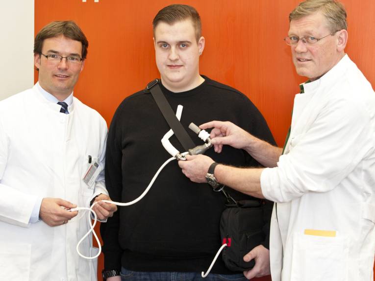 Zwei Männer in weißen Kitteln mit einem jungen Mann, der ein Gerät trägt