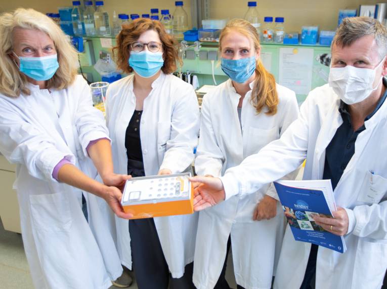 Drei Frauen und ein Mann in weißen Kitteln und mit Nasen-Mund-Schutz-Masken in einem Labor halten etwas in die Kamera.