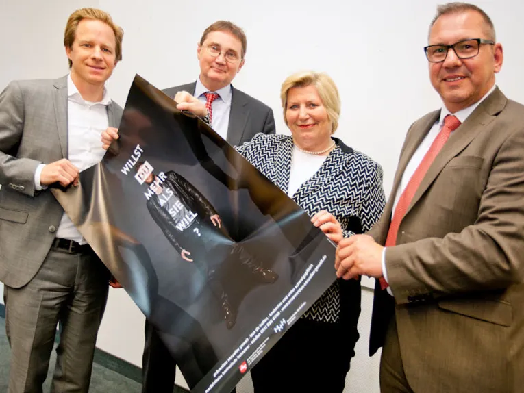 Eine Frau und drei Männer halten ein Plakat in den Händen mit der Aufschrift "Willst Du mehr als sie will?".