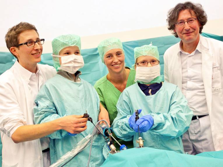 Zwei Männer in weißem Kittel, eine OP-Schwester und zwei Kinder im OP-Saal.