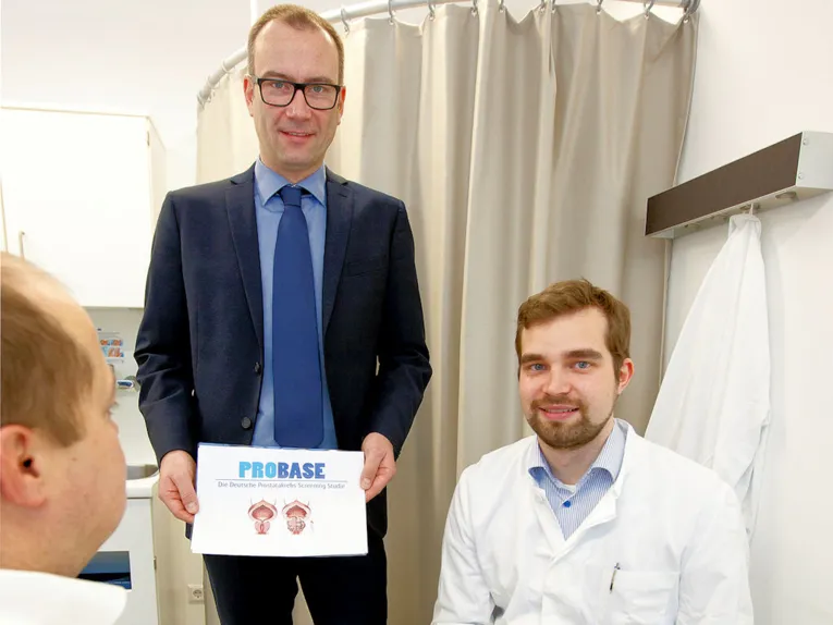 Ein Mann im Anzug stehend mit einem Schild mit der Aufschrift "PROBASE" haltend und ein sitzender Mann in einem weißen Kittel.