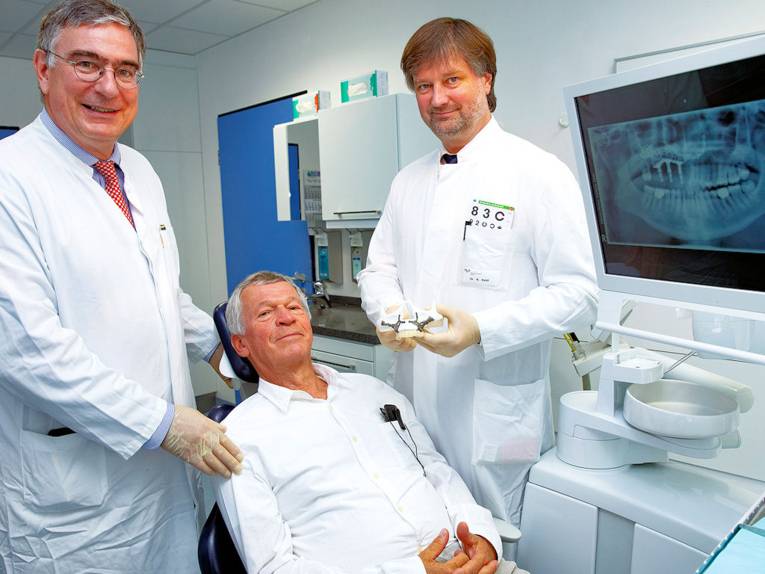 Ein Mann sitzt auf einem Behandlungsstuhl, zwei Männer stehen neben ihm, im Hintergrund ein Röntgenbild.