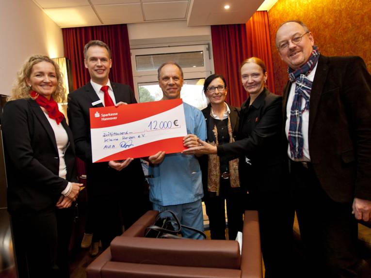 Drei Frauen und drei Männer halten einen symbolischen rot-weißen Check über 12.000 € in die Kamera.