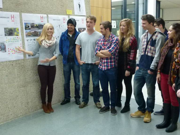 Eine junge Frau zeigt auf ein Plakat, eine Gruppe junger Menschen schaut beziehungsweise hört ihr zu