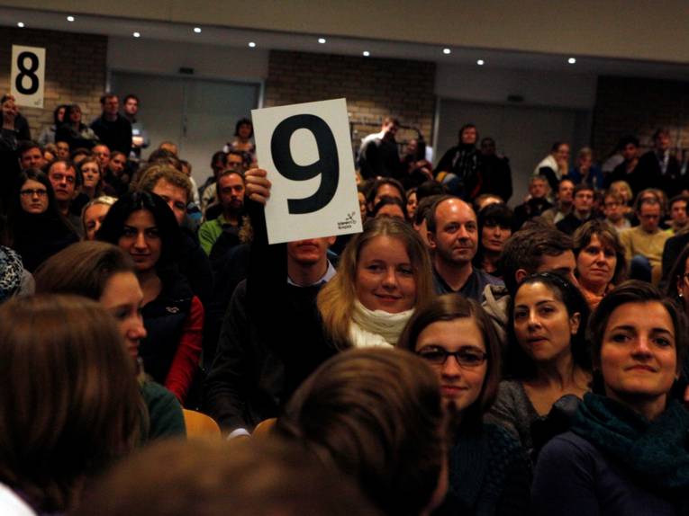 Viele Menschen in einem Raum, einige halten mit Zahlen beschriftete Karten in die Höhe.