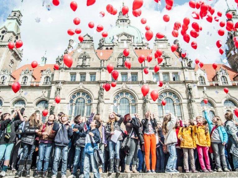 Viele Kinder stehen vor einem alten Gebäude und lassen rote Luftballons fliegen.