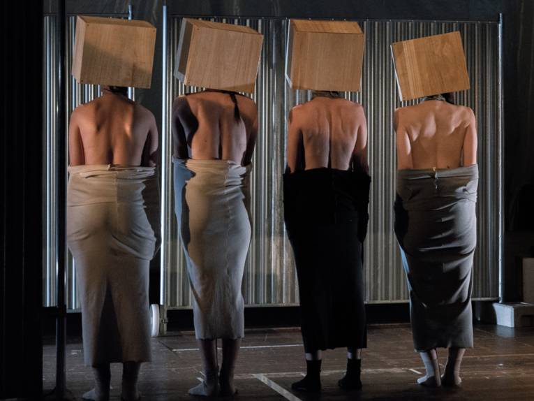 Vier Frauen stehen mit dem Rücken zum Publikum auf einer Bühne und tragen Kartons über ihren Köpfen.