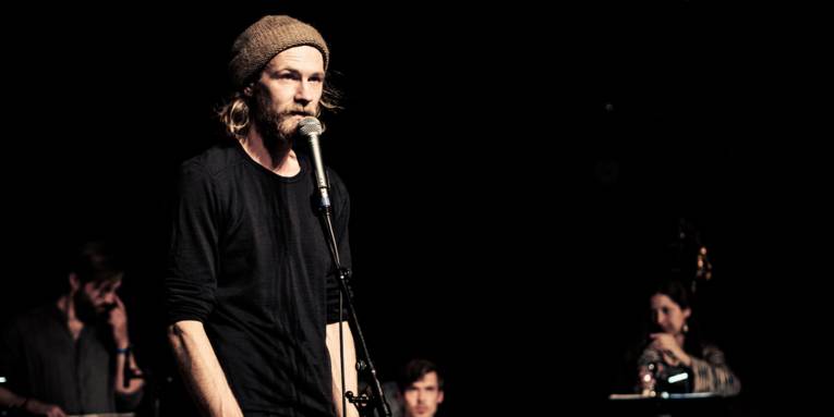 Ein bärtiger Mann mit Wollmütze an einem Mikrophon auf einer Bühne, im Hintergrund drei weitere Musiker.