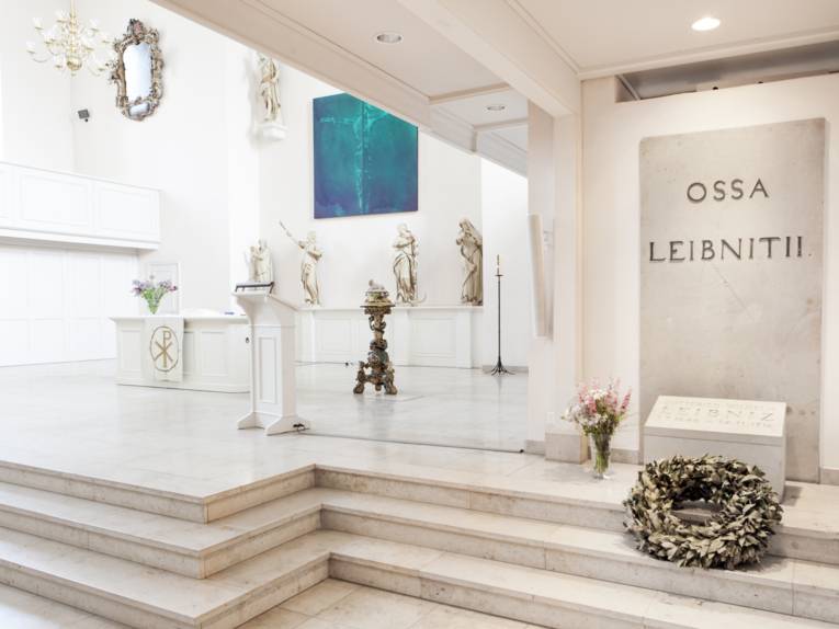 Grabstätte in einem Raum. An der Wand eine Steinplatte, auf der "OSSA LEIBNITII" steht. Davor liegt auf zwei Treppenstufen ein Kranz, links daneben steht eine Vase mit Blumen. Linke Hälfte des Bildes öffnet sich der Raum und man sieht einen Altar mit Statuen sowie eine Rednerpult. 