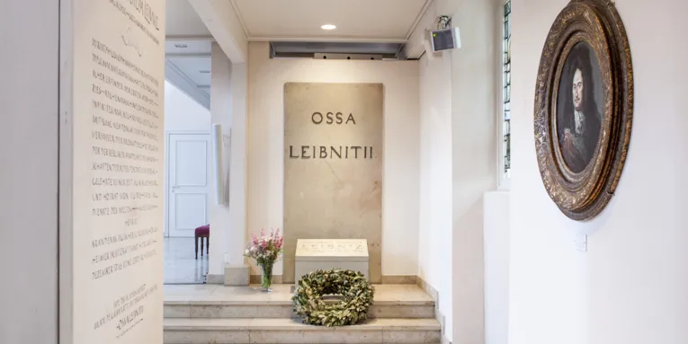 Grabstätte in einem Raum. An der Wand eine Steinplatte, auf der "OSSA LEIBNITII" steht. Davor liegt auf zwei Treppenstufen ein Kranz, links daneben steht eine Vase mit Blumen. Links hinten ist ein Durchgang erkennbar, links vorne eine weitere, beschriftete Steinplatte an einer Wand. In der rechten Wand ist ein rundes Fenster eingelassen, daneben hängt ein Gemälde. 
