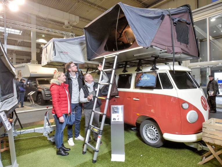 Eine Frau und zwei Männer vor einem VW Bulli Oldtimer mit Dachzelt in einer Messehalle