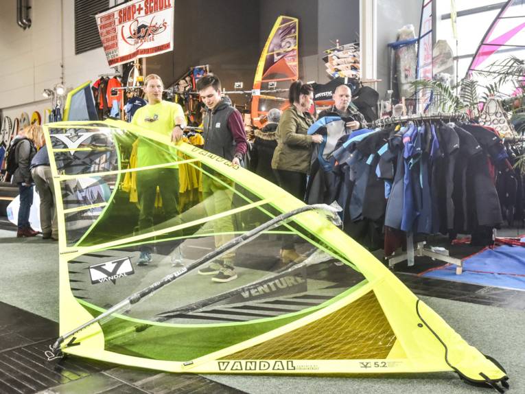 Zwei Männer halten in einer Messehalle des Segel eines Windsurfbrettes.