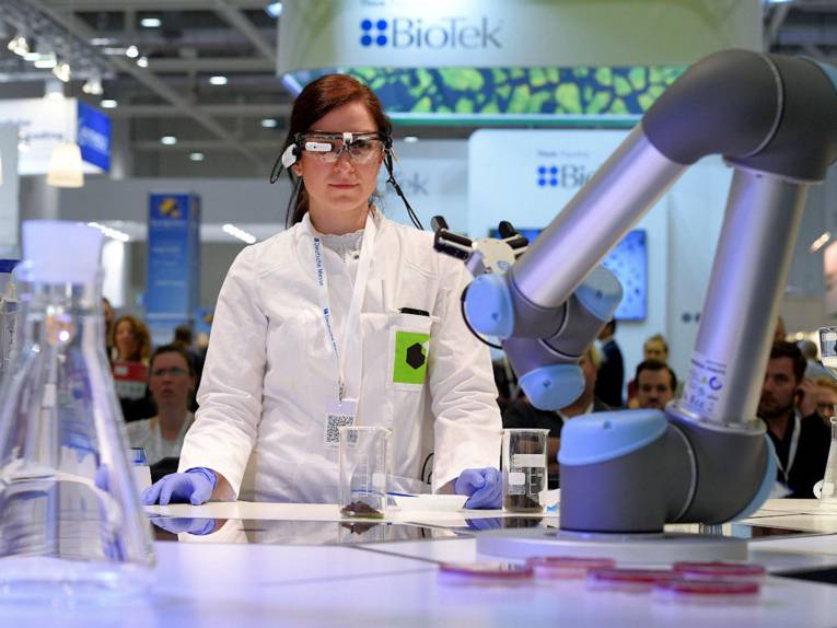 Frau in weißem Kittel mit Schutzbrille und Industrieroboter