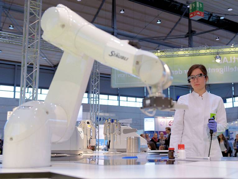 Frau in weißem Kittel mit Schutzbrille und Industrieroboter