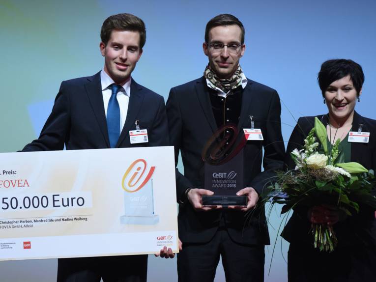 Zwei Männer und eine Frau auf einer Bühne. Der Mann links hält einen überdimensionalen Scheck, der Mann in der Mitte einen kleinen, flachen Gegenstand, die Frau rechts einen Blumenstrauß.