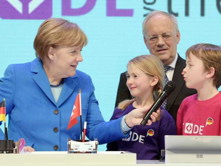 Eine Frau hält zwei Kindern ein Mikrophon hin, im Hintergrund ein Mann.