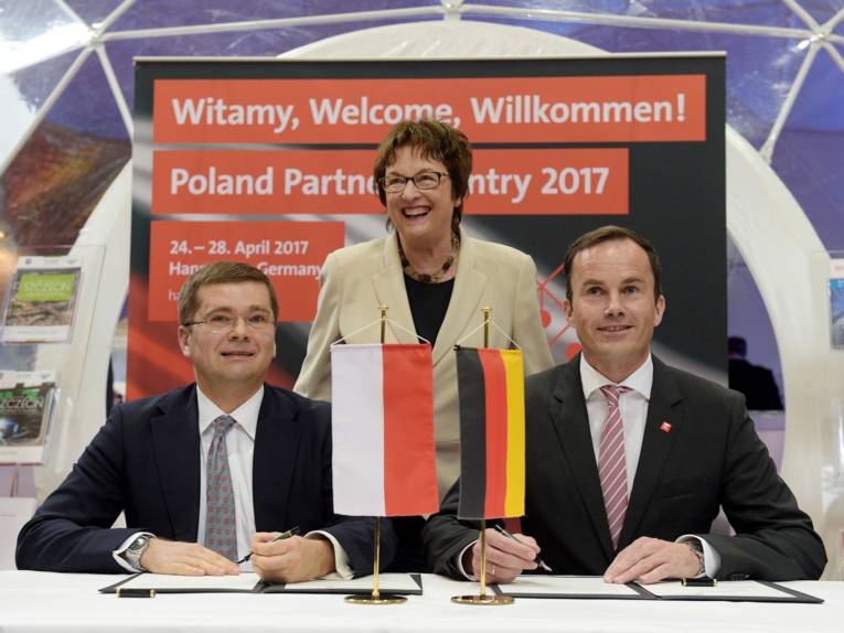 Zwei sitzende Männer unterschreiben etwas, dahinter eine stehende Frau.