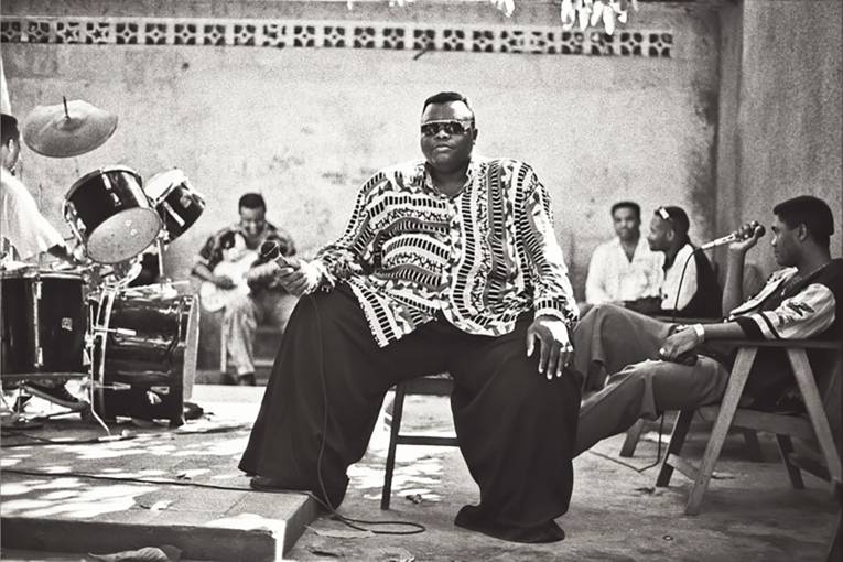 Sechs dunkelhäutige Männer sitzen auf Stühlen auf einer Bühne, teils mit Instrumenten, teils mit Mikrofon.