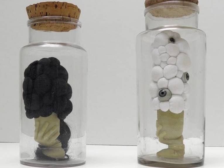 Zwei gefüllte Glasbehälter. Der linke enthält eine schwarze Skulptur, an der man eine Hand und Haare erkennt. Der rechte enthält eine Skulptur, die an Hirnwindungen mit darin platzierten Augäpfeln erinnert.