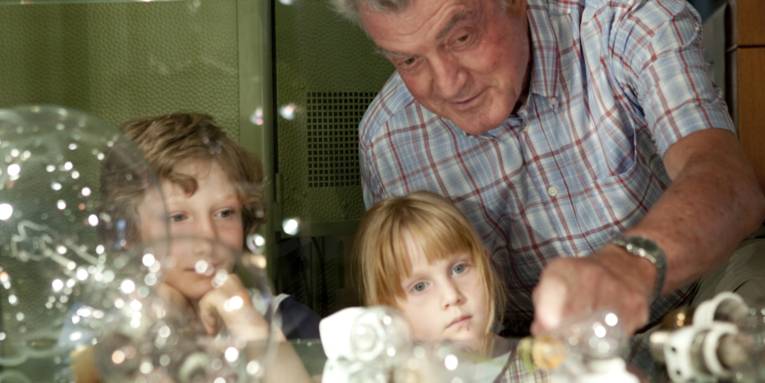 Ein älterer Mann und Kinder stehen vor einem Glasregal mit Glühbirnen.