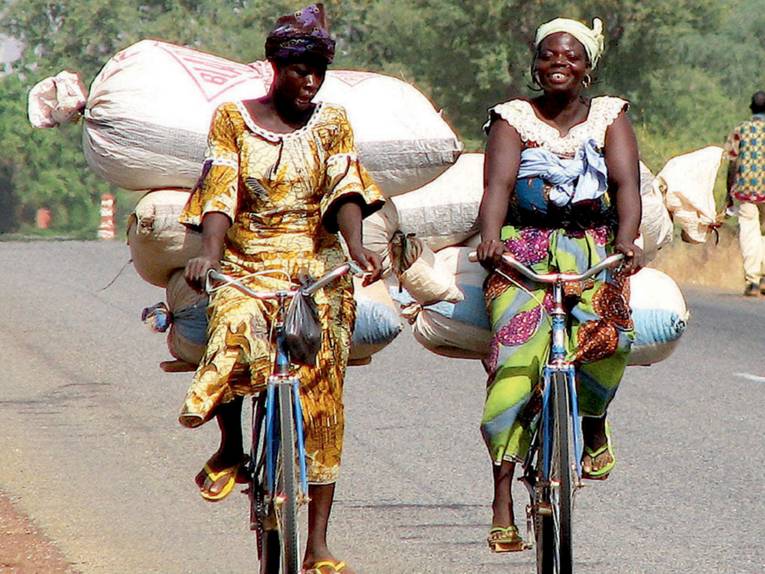 Zwei dunkelhäutige Frauen fahren auf einer Landstraße mit dem Fahrrad, beide Räder sind mit Reissäcken oder ähnlichem hoch beladen.